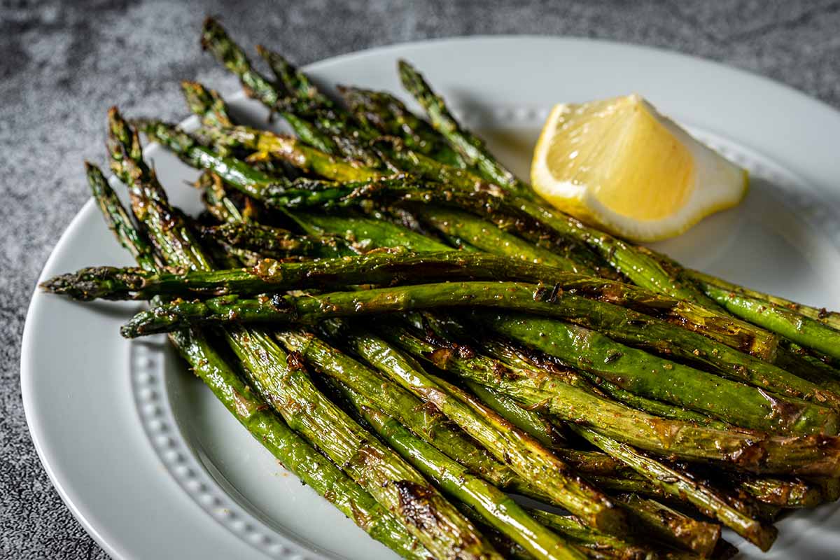A close up horizontal image of grilled asparagus set on a white serving dish with a slice of lemon.
