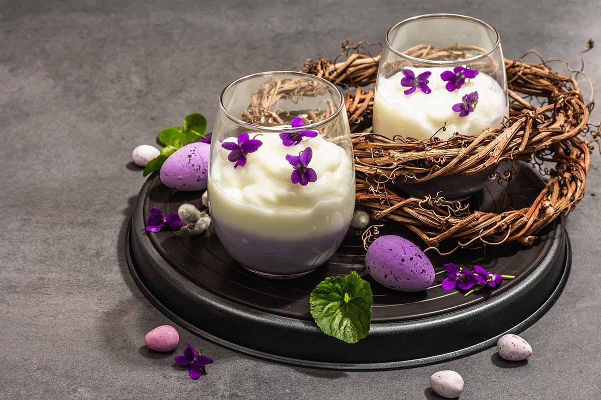 A horizontal photo of two cups of custard desserts topped with whipped cream and purple blossoms on top.