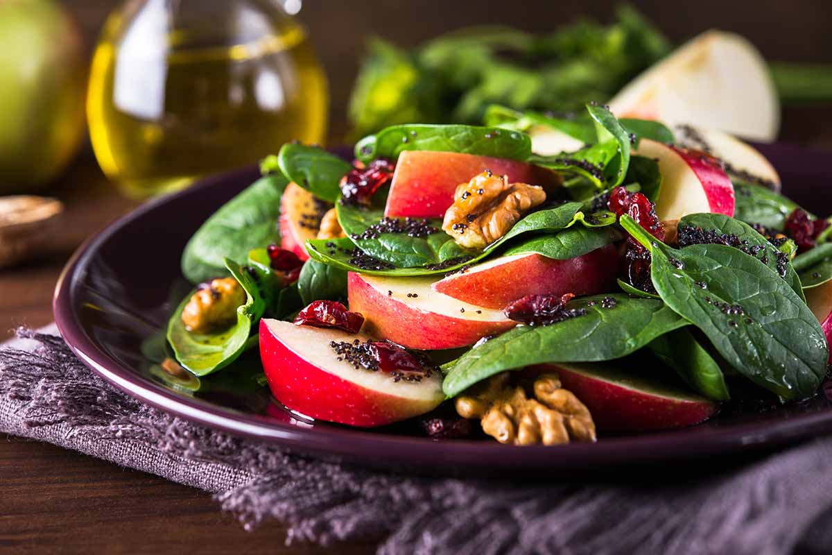 A horizontal photo of a fresh spinach salad with apples and walnuts on a dark plate atop a placemat and table.