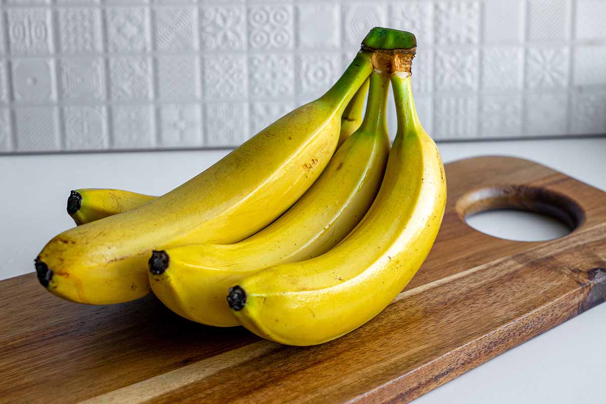A close up horizontal image of a hunch of bananas set on a wooden chopping board with a background of ceramic tiles.
