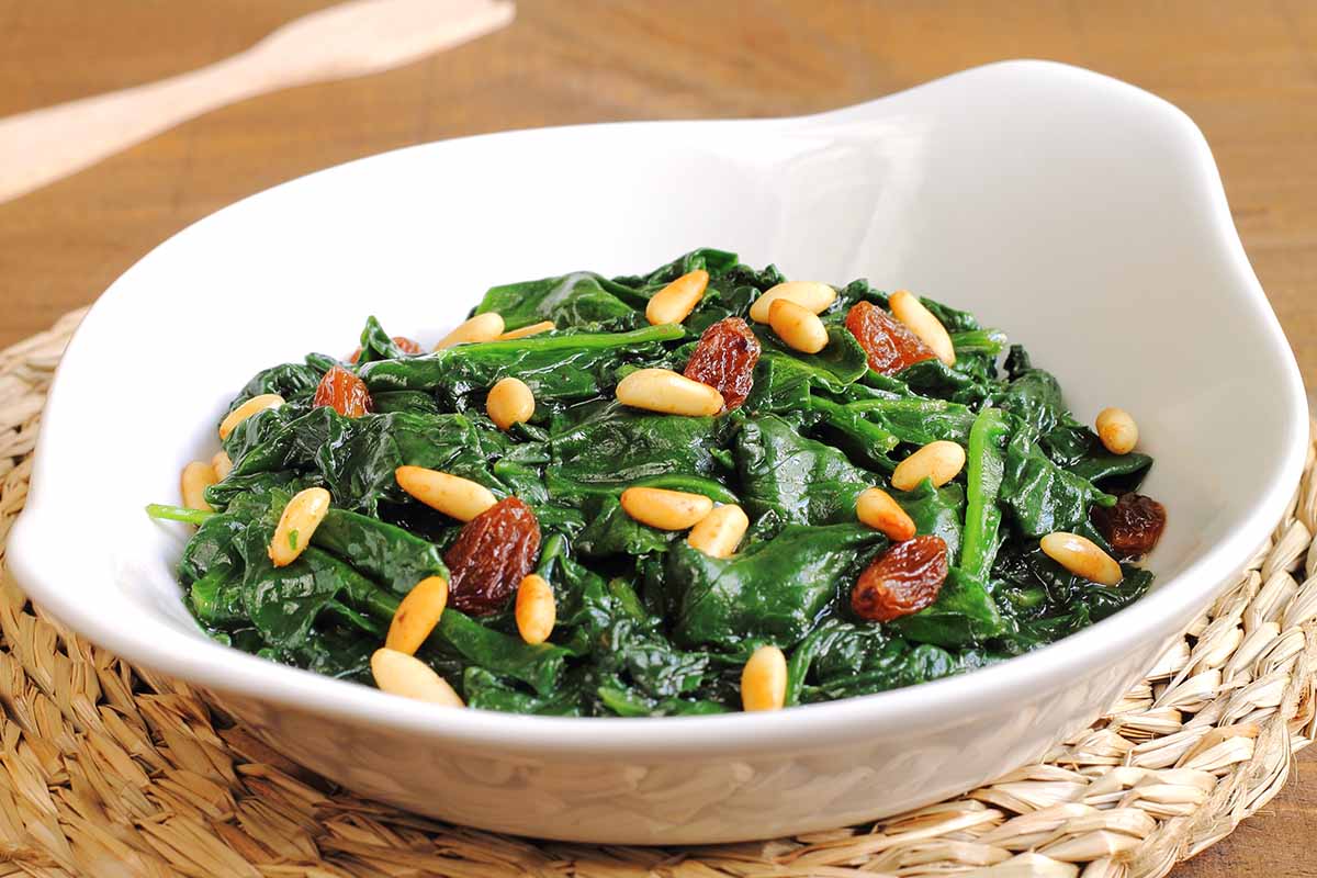 A horizontal photo of sauteed spinach with raisins and pine nuts in a white pottery bowl on a wicker placemat.