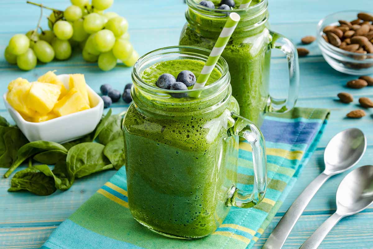A horizontal photo of two spinach smoothies in mason jar mugs surrounded by the fresh ingredients used in the smoothies.