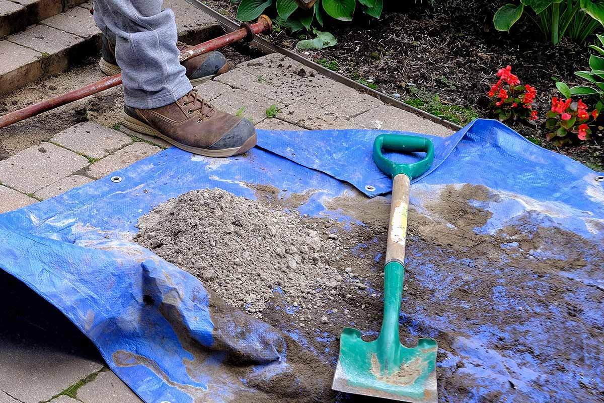 A close up horizontal image of a gardener working on a garden border with a tarp and spade to the bottom of the frame.