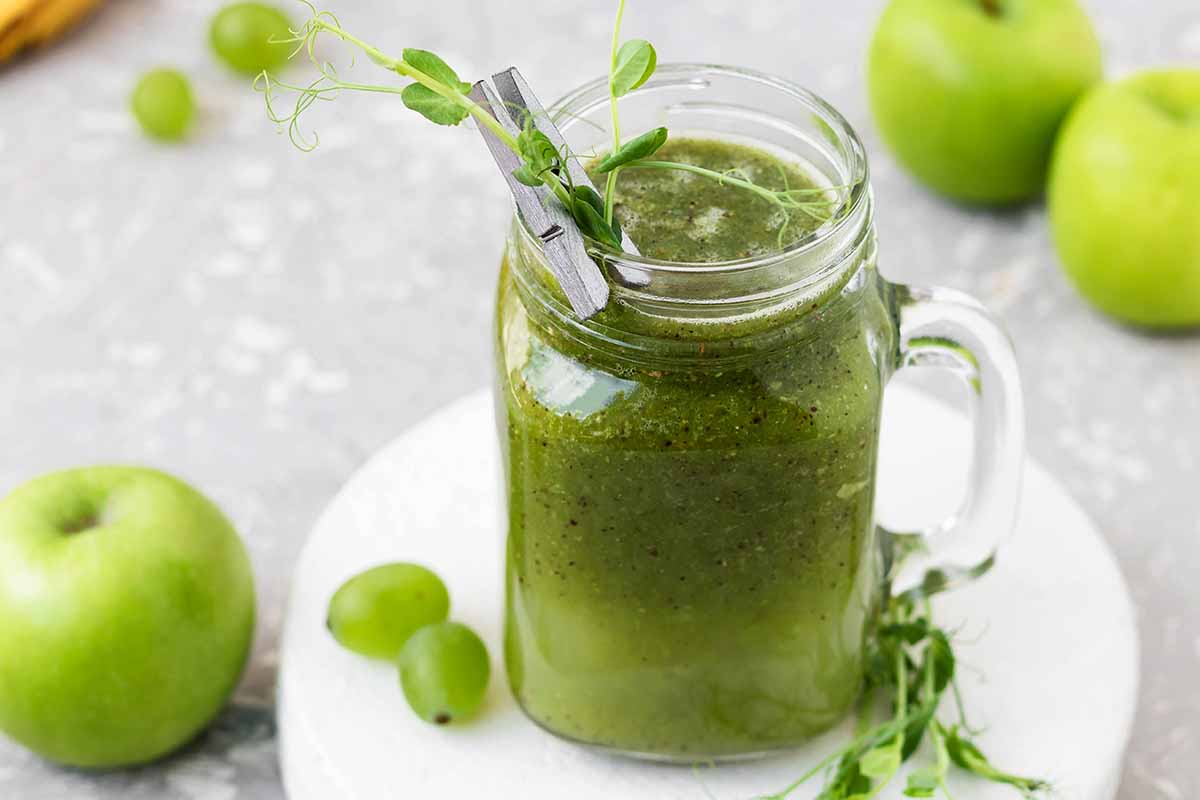A horizontal photo of pea shoots in a green smoothie in a glass mason jar. Pea shoots, grapes and green apples are lying next to the smoothie on a table.