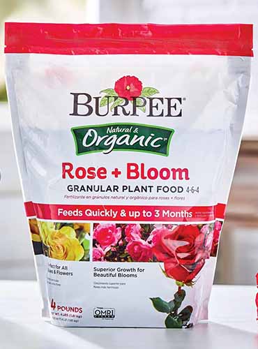 A close up of a bag of Rose + Bloom granular plant food set on a white surface indoors.