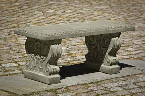 A close up horizontal image of the Finkel Cast Stone Garden Bench set on a concrete patio.