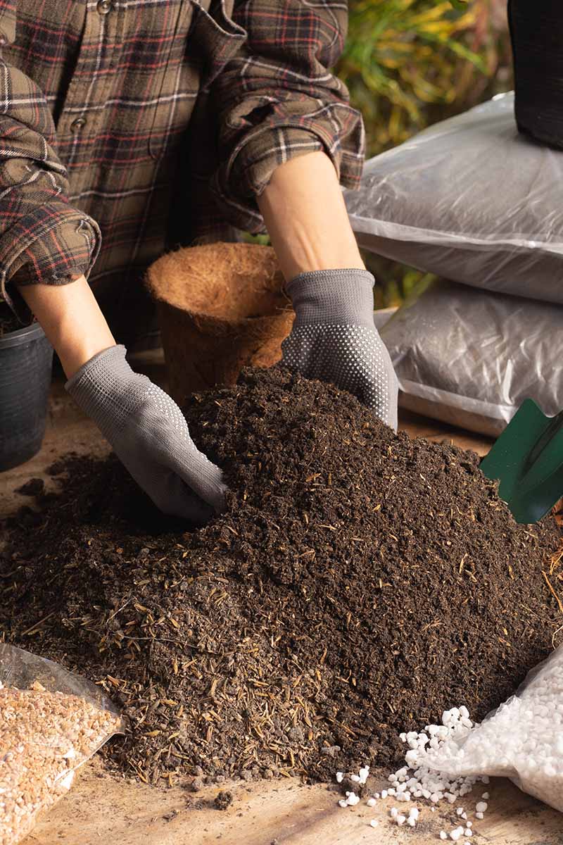 A close up vertical image of a gardener wearing gloves to mix soil for planting in pots.