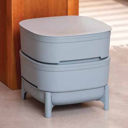 A close up of a light gray plastic Wow Wow Farm Composter set on the ground indoors.