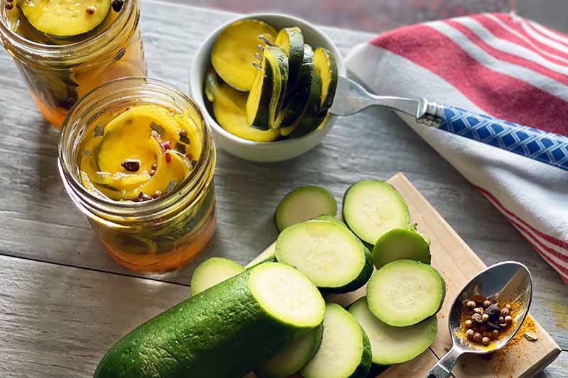 A horizontal photo of a table with sliced zucchini, and jars of bread and butter pickles.