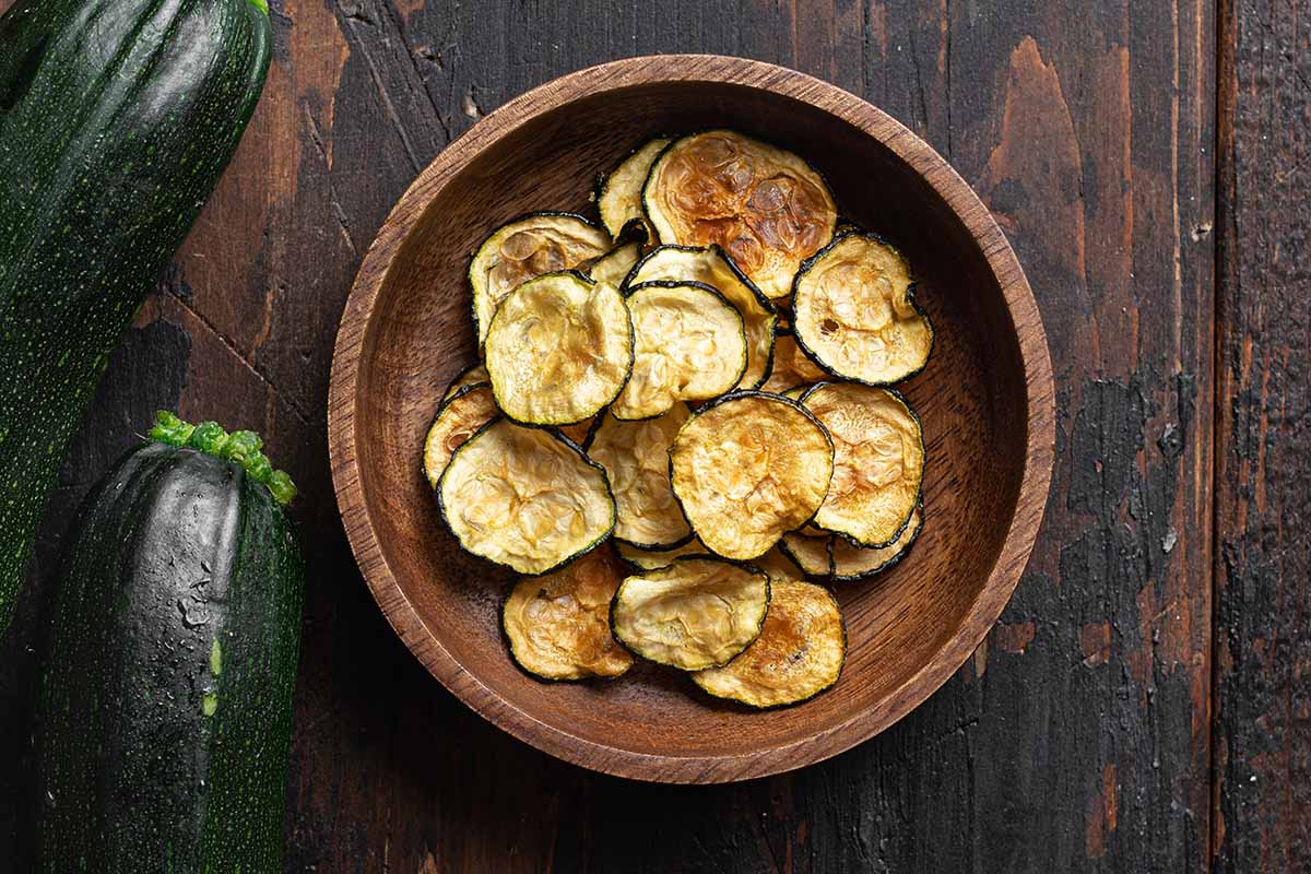 A horizontal photo shot from above of a wooden bowl filled with dehydrated zucchini chips on a wooden table.