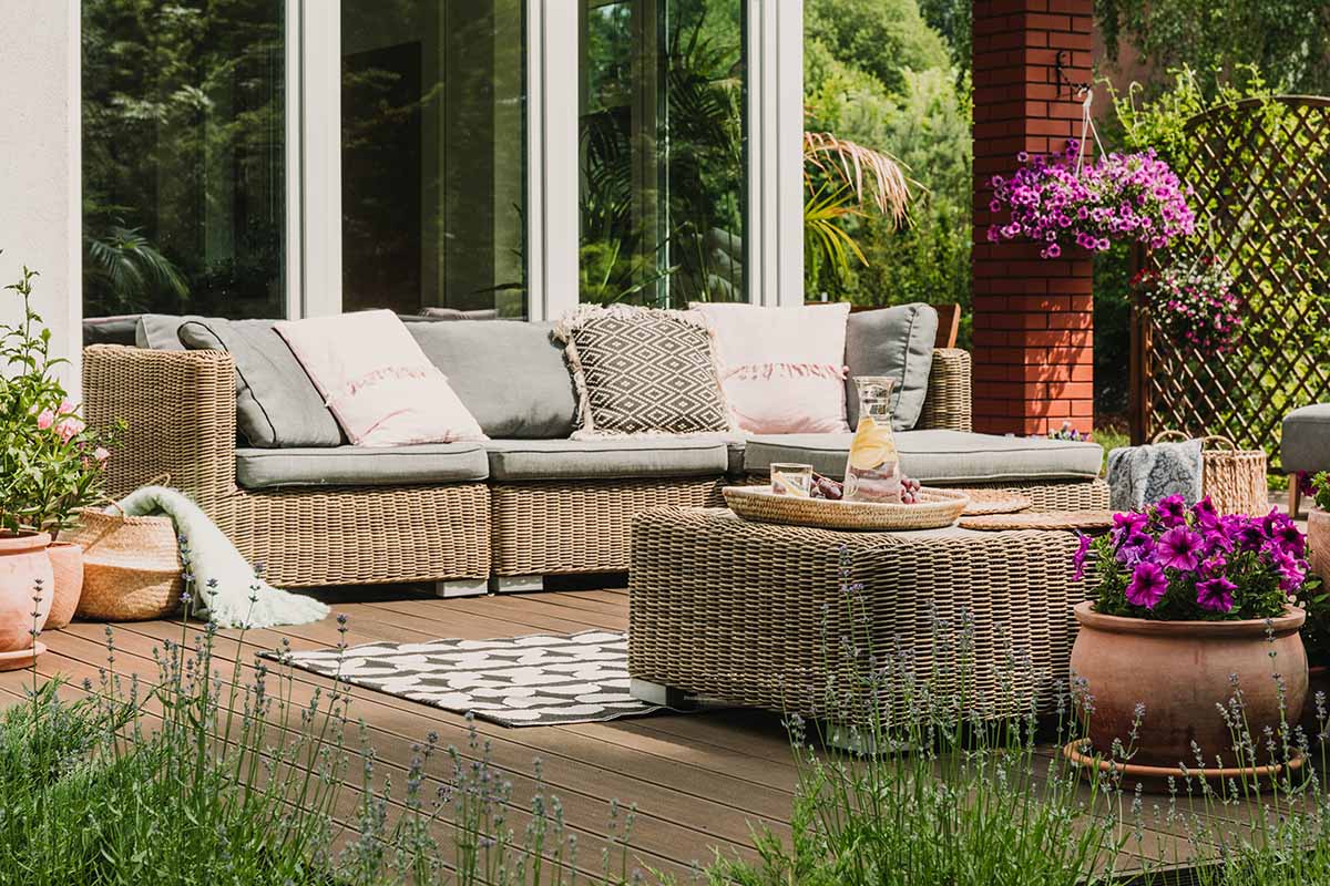A horizontal image of a deck scene with wicker furniture, rugs, and a variety of plants.