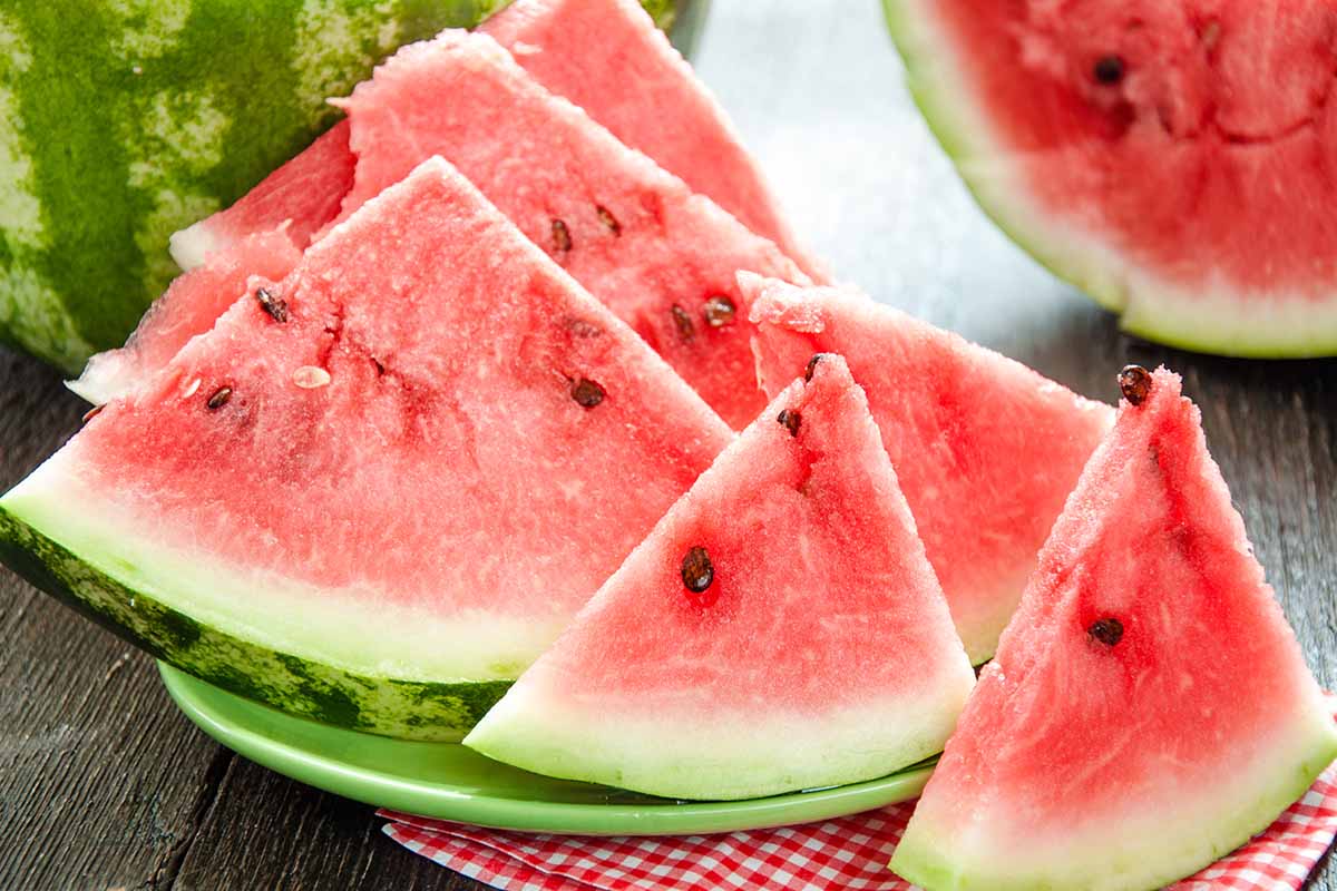 A close up of slices of watermelon set on a green plate on a wooden surface with fruit in soft focus in the background.