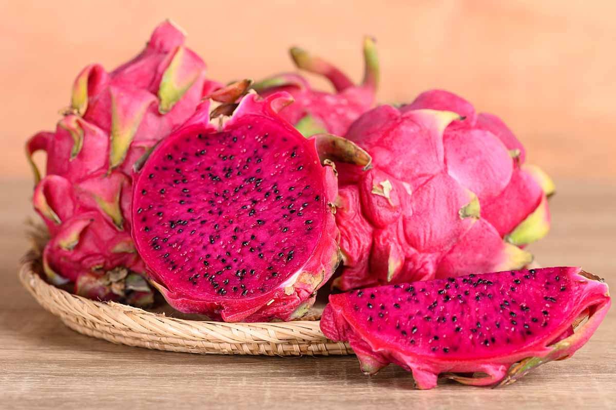 A close up horizontal image of red dragon fruit (pitaya) whole and sliced in half set on a wicker tray on a wooden surface.