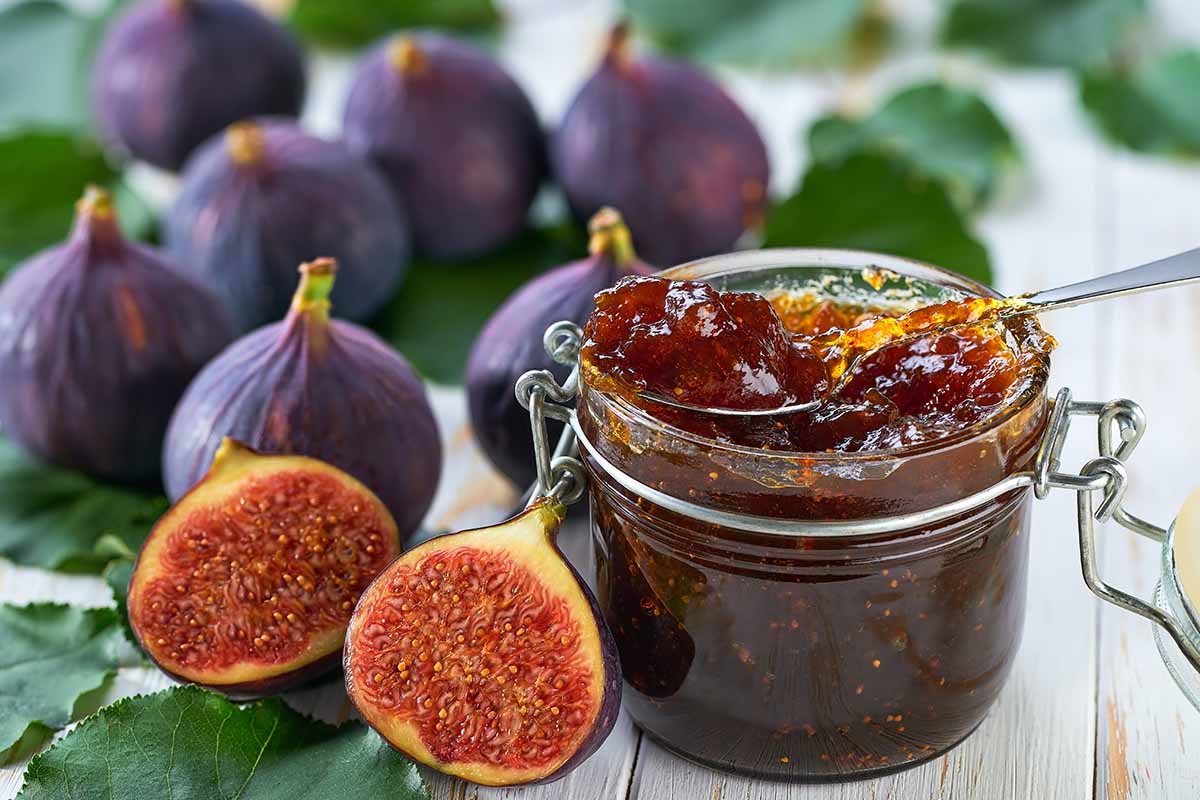 A close up horizontal image of a glass jar filled with jam and a variety of whole and sliced figs, set on a wooden surface, decorated with foliage.