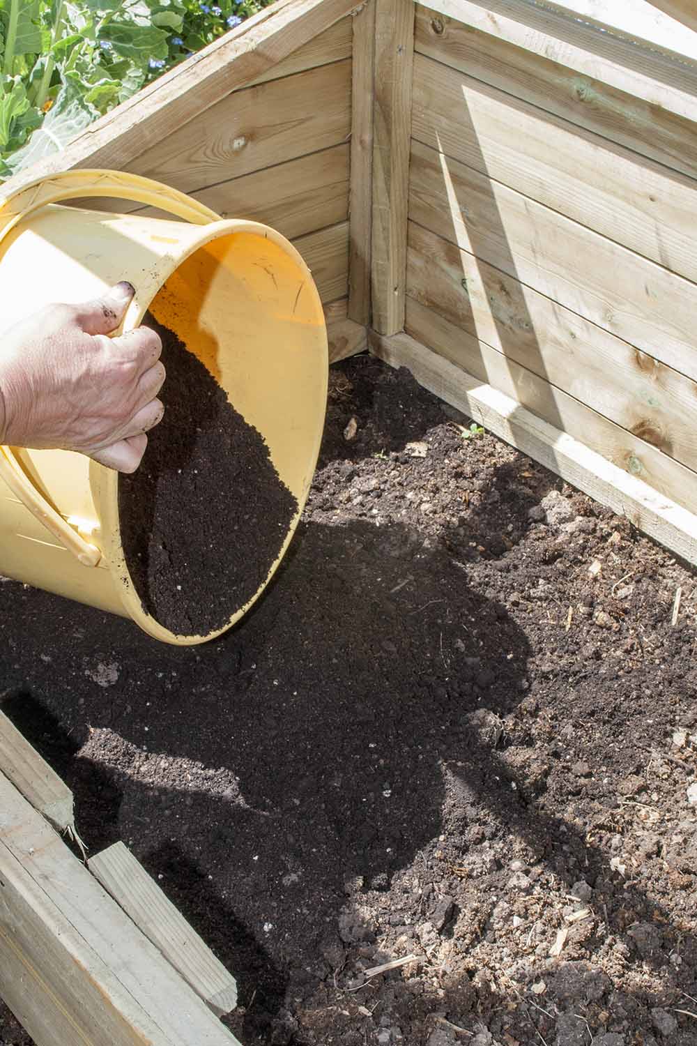 A close up vertical image of a hand from the left of the frame emptying soil into a wooden raised bed.