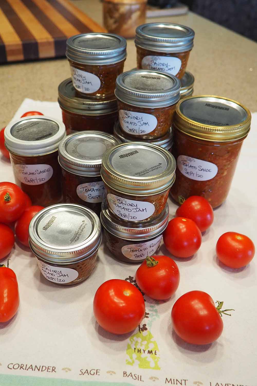 A close up vertical image of a variety of canned sauces made from a fresh tomato harvest from the garden, with ripe fruits scattered around.