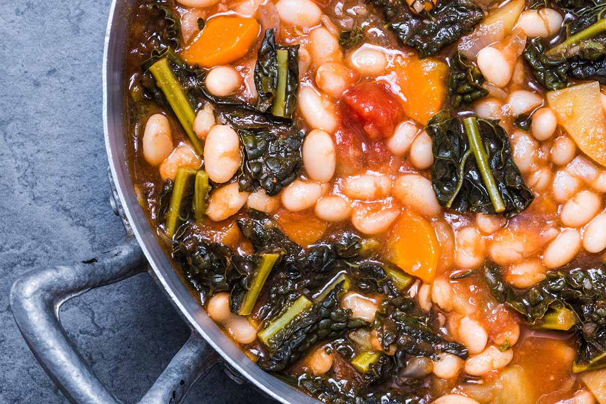 A close up top down horizontal image of a metal saucepan of a beans and greens dish freshly cooked set on a gray surface.