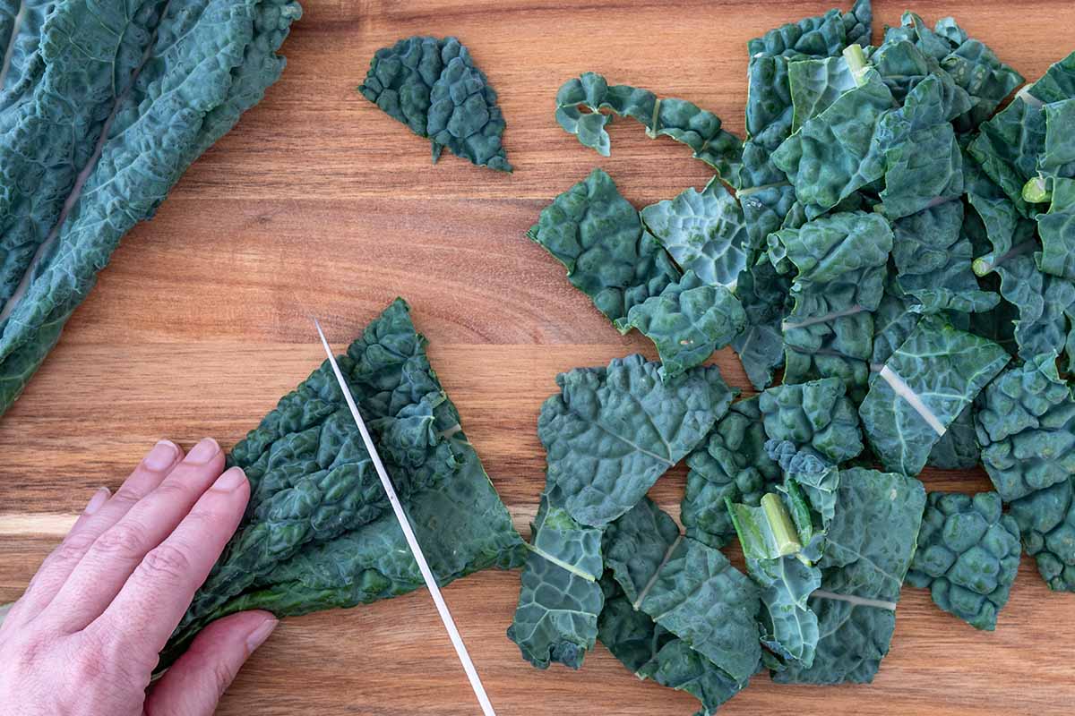 A close up horizontal image of a hand to the left of the frame chopping leafy greens into small chunks on a wooden surface for cooking.
